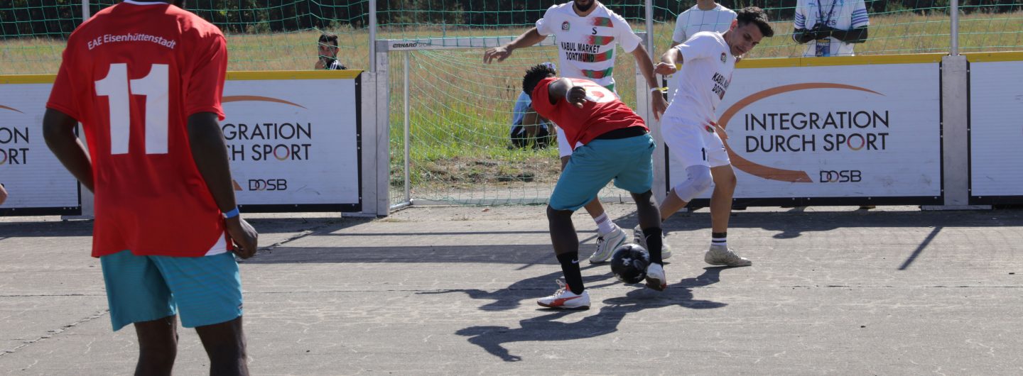 Ein Fußballfeld. Links steht ein Spieler. Auf seinem Trikot steht "EAE Eisenhüttenstadt". Rechts sind drei weitere Spieler zu sehen, die um den Ball kämpfen.