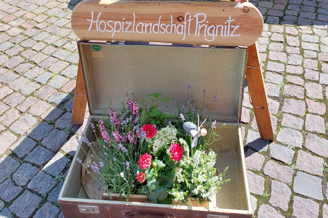 Ein Blumenstrauß in einem Koffer. Darüber ein Holzschild: Hospizlandschaft Prignitz.