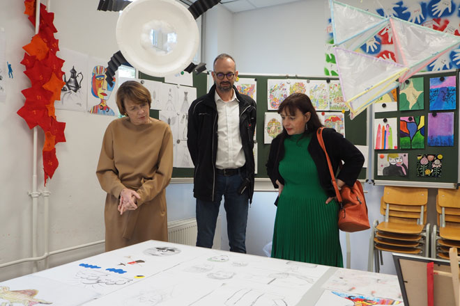 In einem Kunstraum stehen links Elke Büdenbender, in der Mitte Christian Seiler und rechts Wiebke Şahin-Schwarzweller und betrachten Kunst auf einem Tisch.