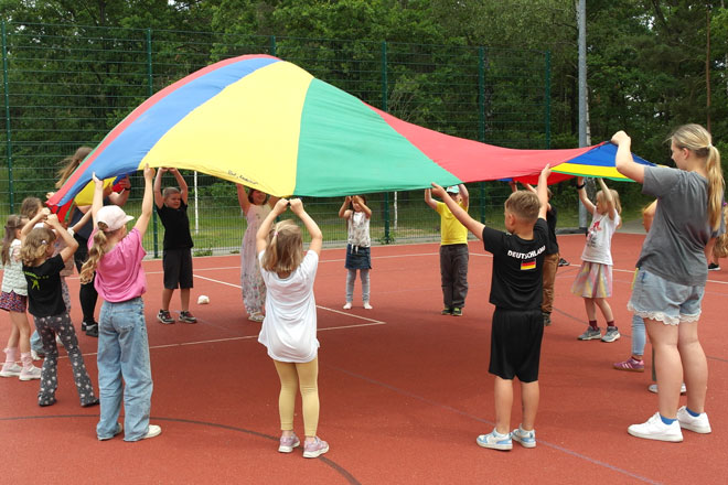 Schulkinder stehen auf einem Sportplatz in einem Kreis und halten ein buntes Schwungtuch in die Luft.