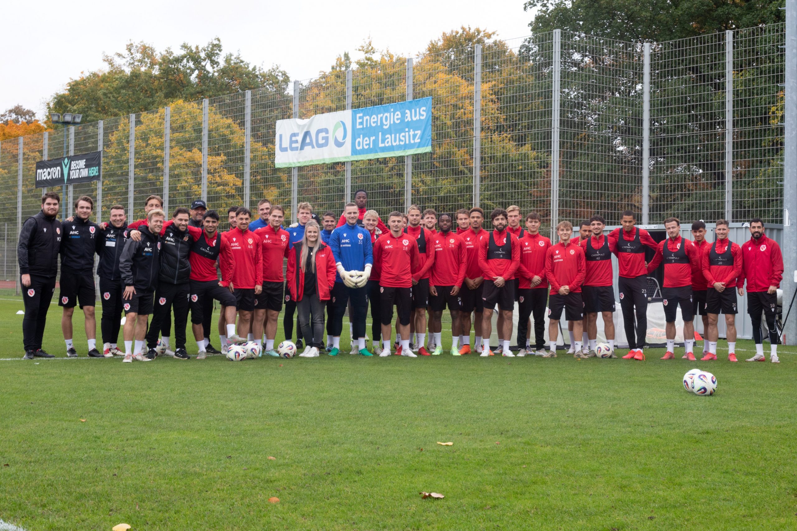 Fußballer des FC Energie Cottbus stehen in Trainingskleidung aufgreiht auf einem Fußballplatz. In ihrer Mitte steht eine Frau mit einer Jacke des DRK.