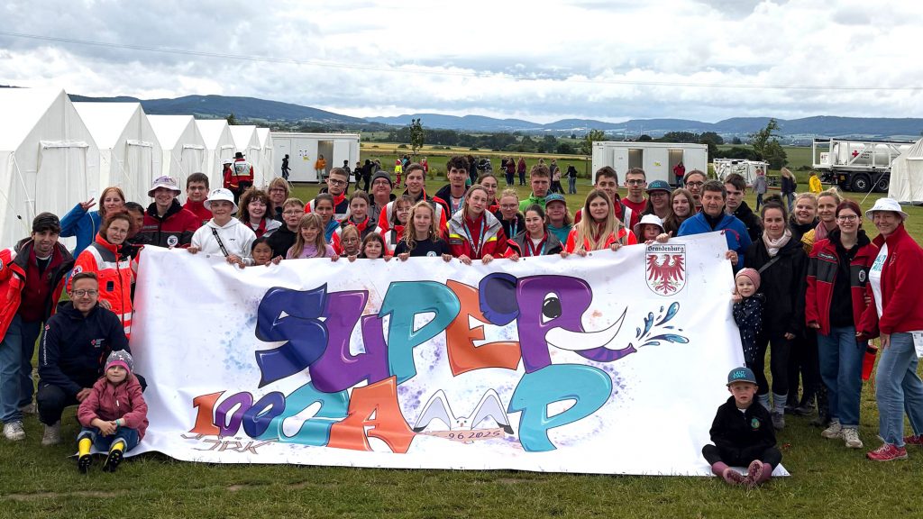 Zu sehen ist ein Gruppebild von rund 40 vorwiegend jungen Menschen, die bei einem Zeltlager eine großes Leintuch halten, auf dem "Supercamp - 100 Jahre JRK" steht.