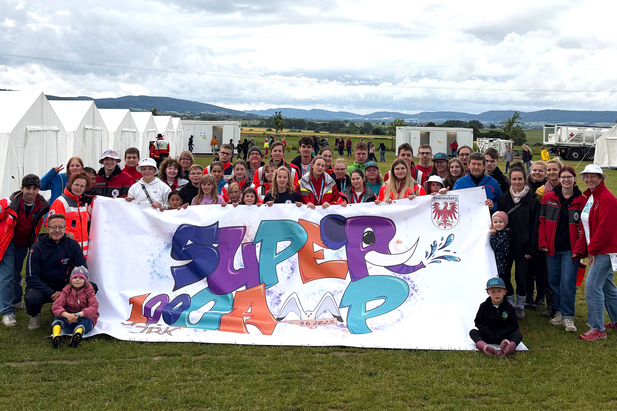 Zu sehen ist ein Gruppebild von rund 40 vorwiegend jungen Menschen, die bei einem Zeltlager eine großes Leintuch halten, auf dem "Supercamp - 100 Jahre JRK" steht.