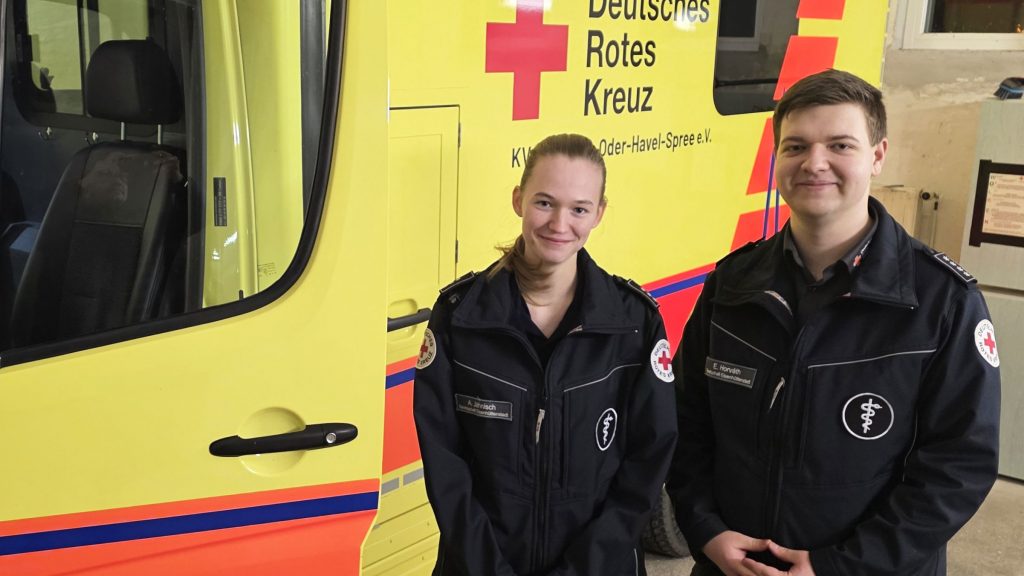 Eine junge Frau und ein junger Mann in Einsatzkleidung stehen vor einem Krankentransportwagen.