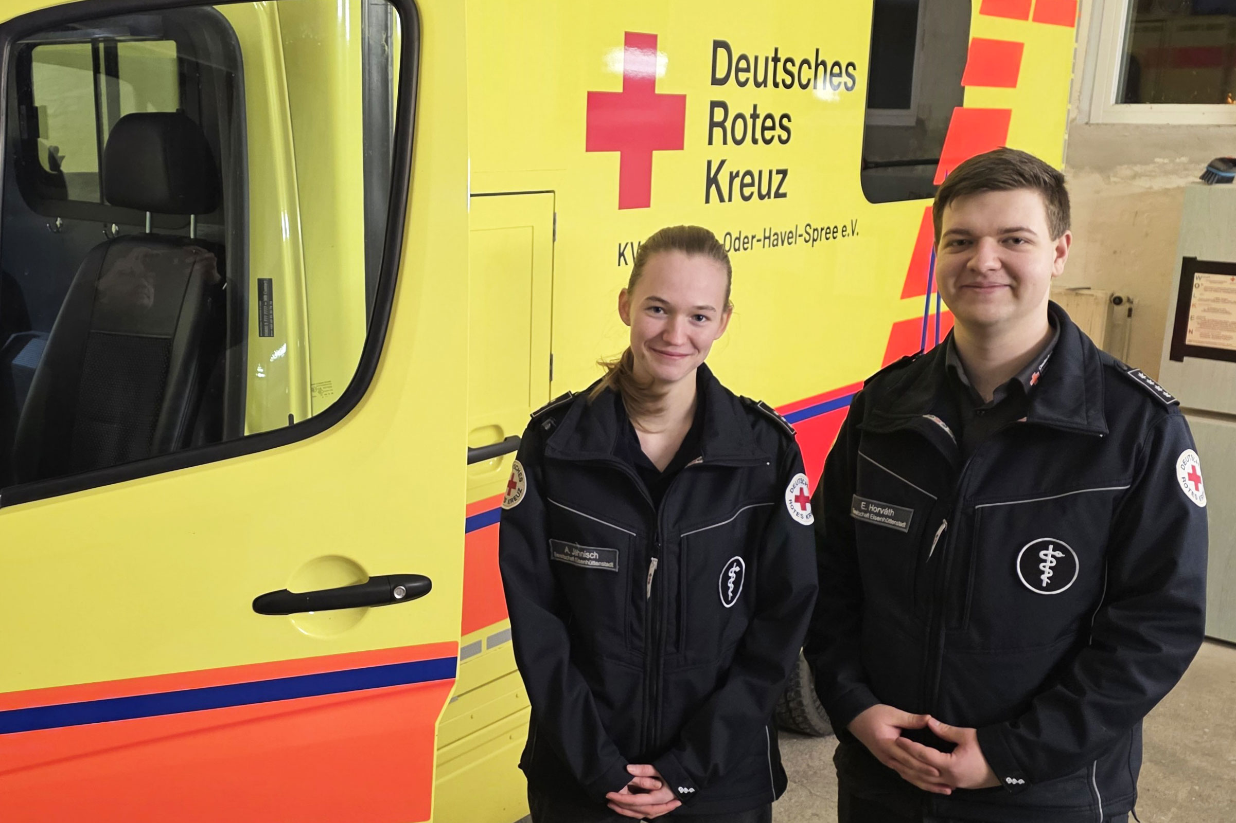 Eine junge Frau und ein junger Mann in Einsatzkleidung stehen vor einem Krankentransportwagen.