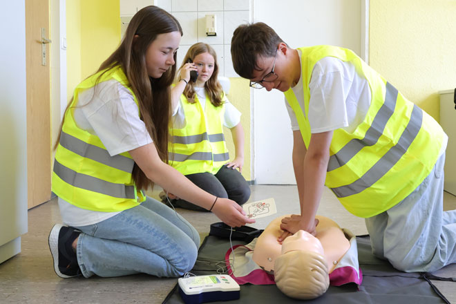 Zwei Jugendliche in gelben Warnwesten üben Wiederbelebung an einer Puppe. Im Hintergrund hält eine dritte Jugendliche in gelber Warnweste ein Handy am Ohr.
