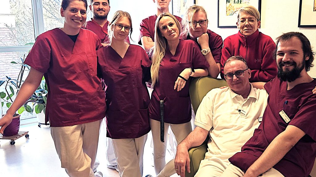 Rund 10 Menschen in Arbeitskleidung einer Pflegeeinrichtung stehen in einer Gruppe zusammen und schauen lächelnd in die Kamera