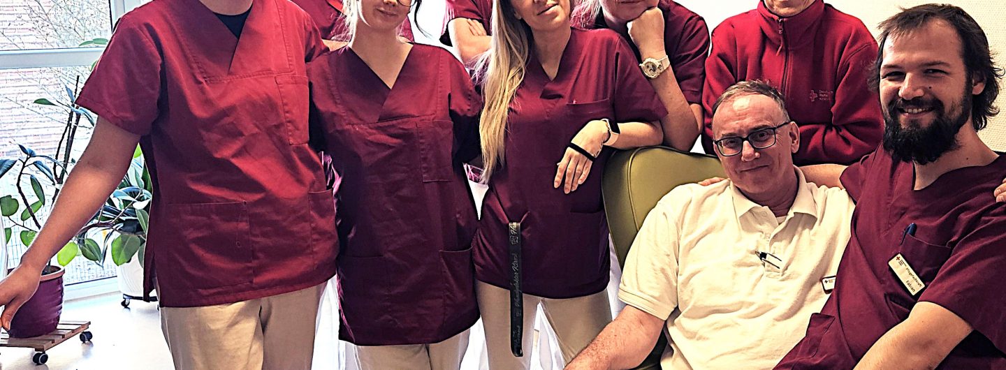 Rund 10 Menschen in Arbeitskleidung einer Pflegeeinrichtung stehen in einer Gruppe zusammen und schauen lächelnd in die Kamera