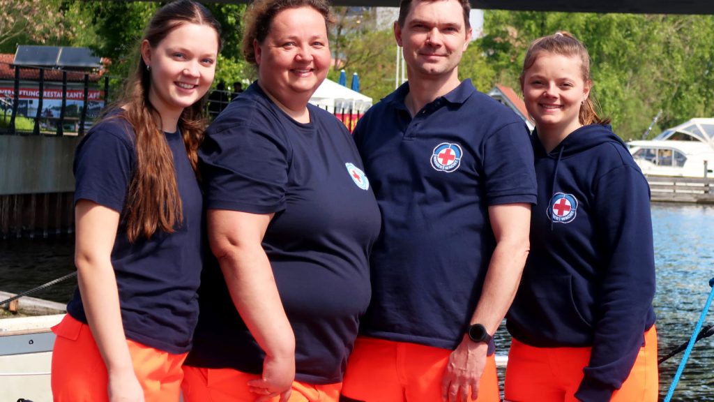 Drei Frauen ud ein Mann stehen in Einsatzkleidung der Wasserwacht des DRK am Wasser.