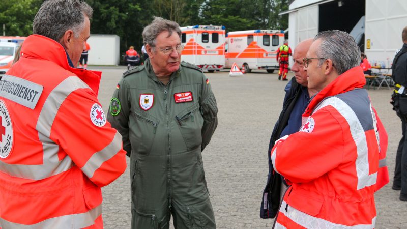 Ein Mann in Uniform der Bundeswehr, zwei Männer in Einsatzkleidung des Roten Kreuzes und ein Mann in Zivilkleidung stehen im Freien zusammen und beraten sich.
