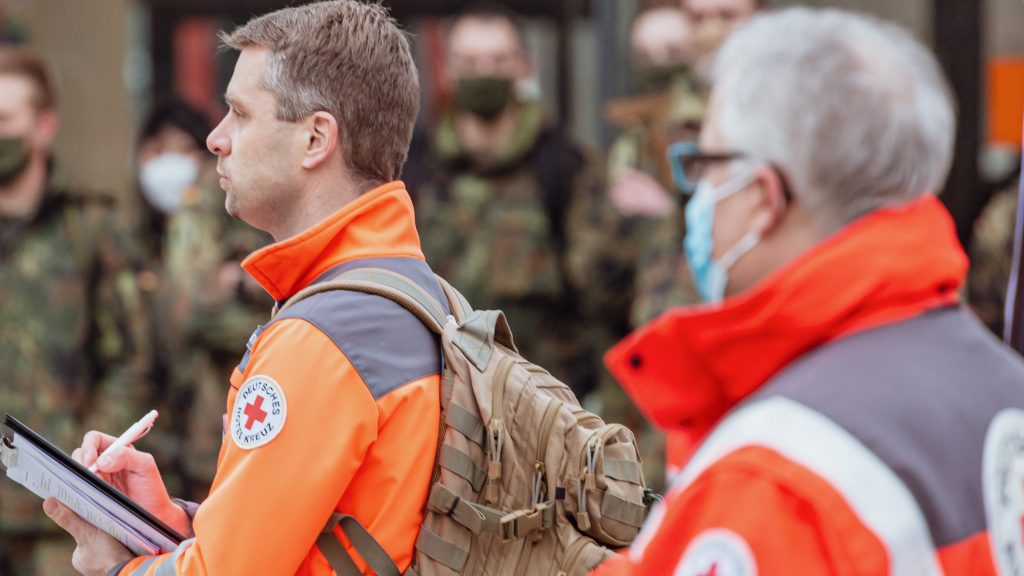 Im Vordergrund stehen zwei Männer in Einsatzkleidung des Deutschen Roten Kreuz, im Hintergrund unscharf Menschen in Uniform der Bundeswehr.