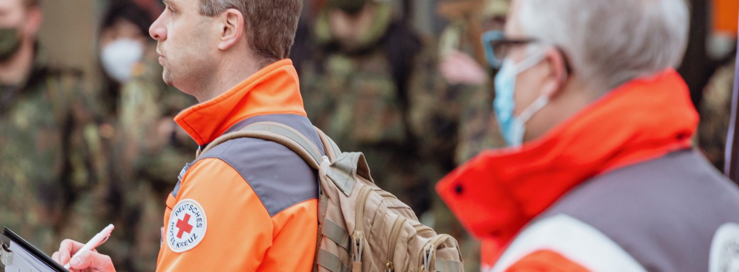 Im Vordergrund stehen zwei Männer in Einsatzkleidung des Deutschen Roten Kreuz, im Hintergrund unscharf Menschen in Uniform der Bundeswehr.