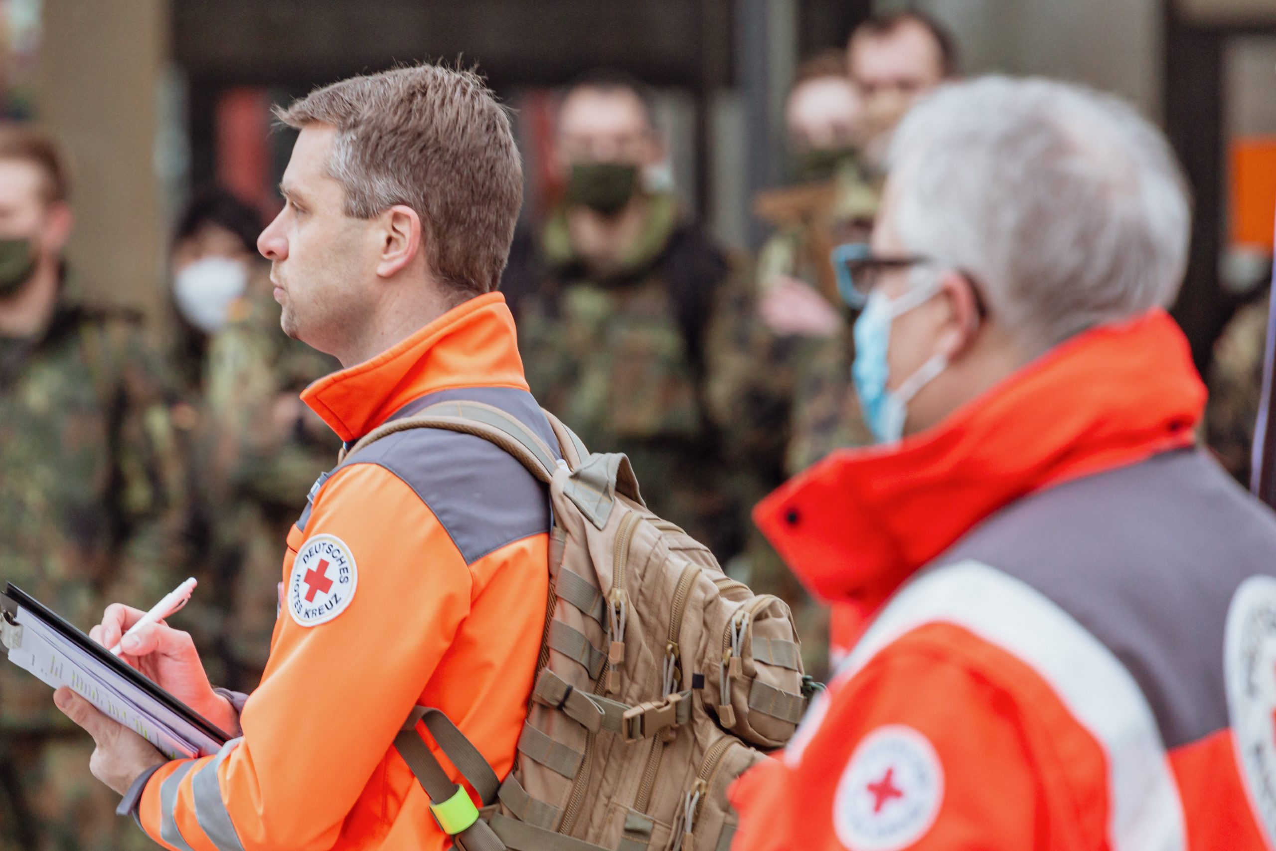Im Vordergrund stehen zwei Männer in Einsatzkleidung des Deutschen Roten Kreuz, im Hintergrund unscharf Menschen in Uniform der Bundeswehr.