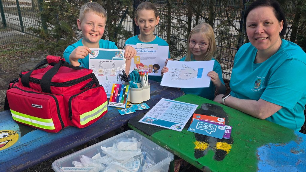 Eine Frau und drei Kinder in T-Shirts des Jugendrotkreuz sitzen an einem bunt bemalten Tisch und halten kindgerechtes Lehrmaterial für Erste Hilfe in die Kamera
