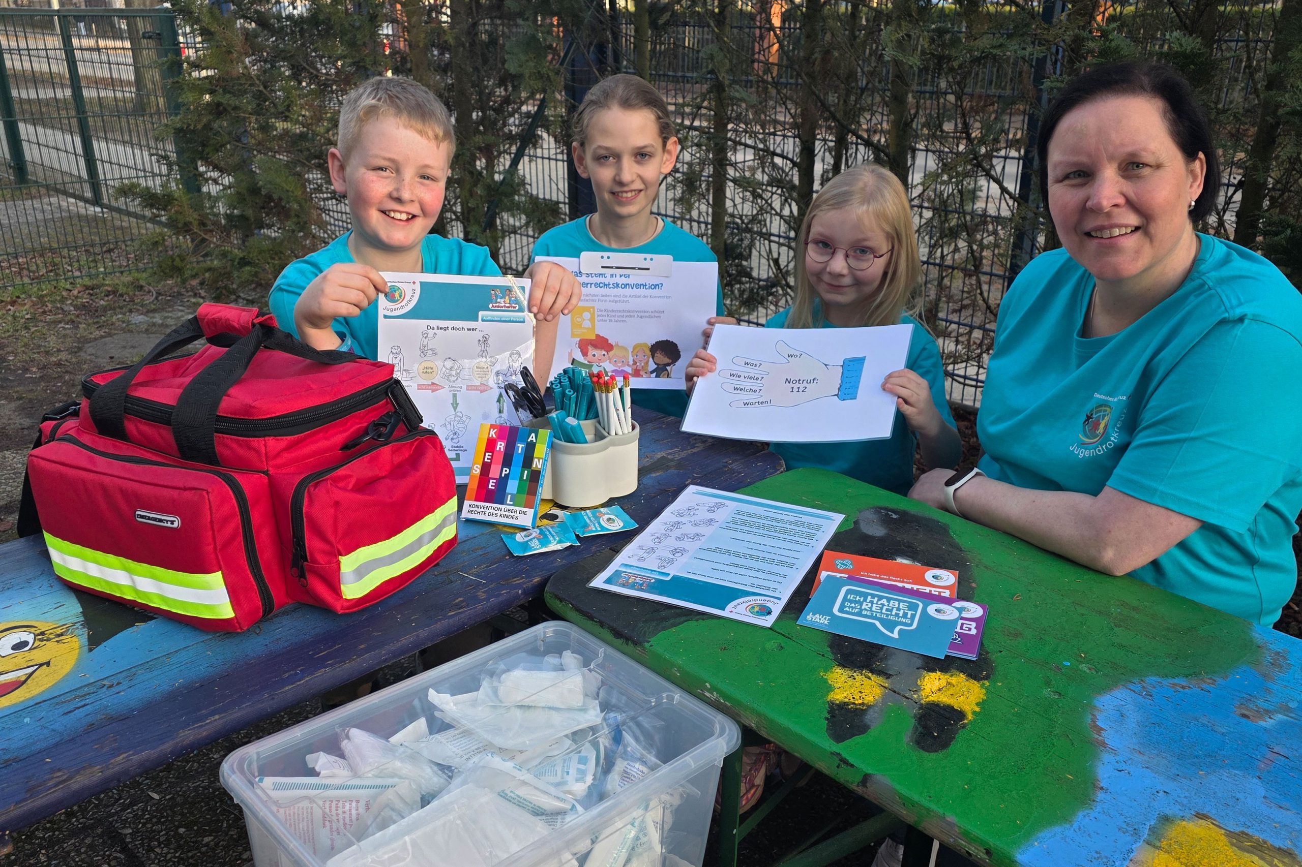 Eine Frau und drei Kinder in T-Shirts des Jugendrotkreuz sitzen an einem bunt bemalten Tisch und halten kindgerechtes Lehrmaterial für Erste Hilfe in die Kamera