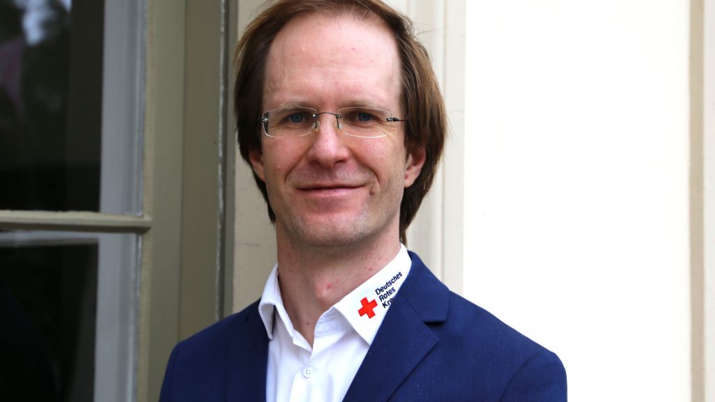 Porträtfoto eines Mannes, der freundlich in die Kamera lächelt. Der Mann trägt ein Jackett und ein Hemd, auf dem ein Logo des Deutschen Roten Kreuz zu sehen ist.