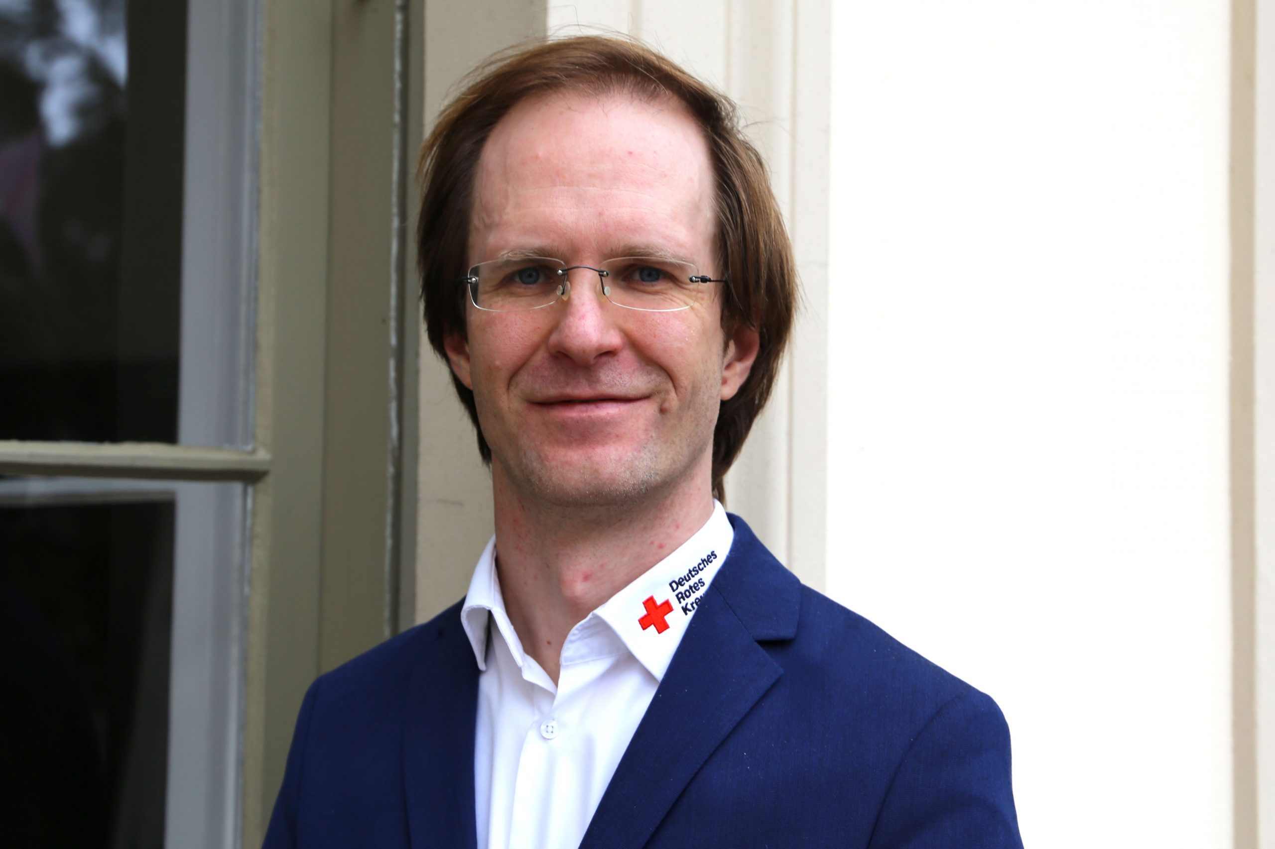 Porträtfoto eines Mannes, der freundlich in die Kamera lächelt. Der Mann trägt ein Jackett und ein Hemd, auf dem ein Logo des Deutschen Roten Kreuz zu sehen ist.