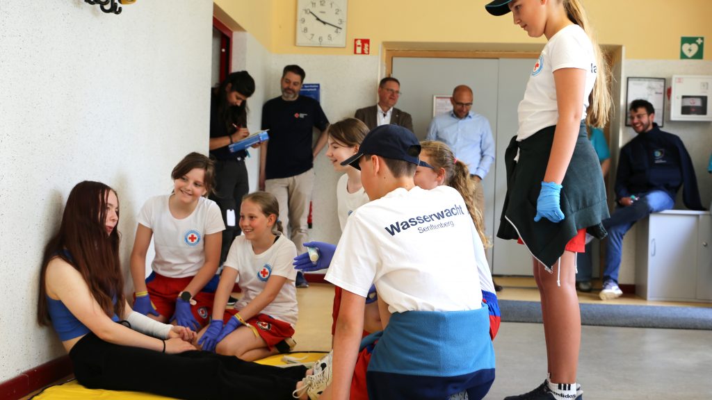 Fünf Kinder in T-Shirts der Wasserwacht des DRK kümmern sich in einer inszenierten Szene um eine Verletzte.