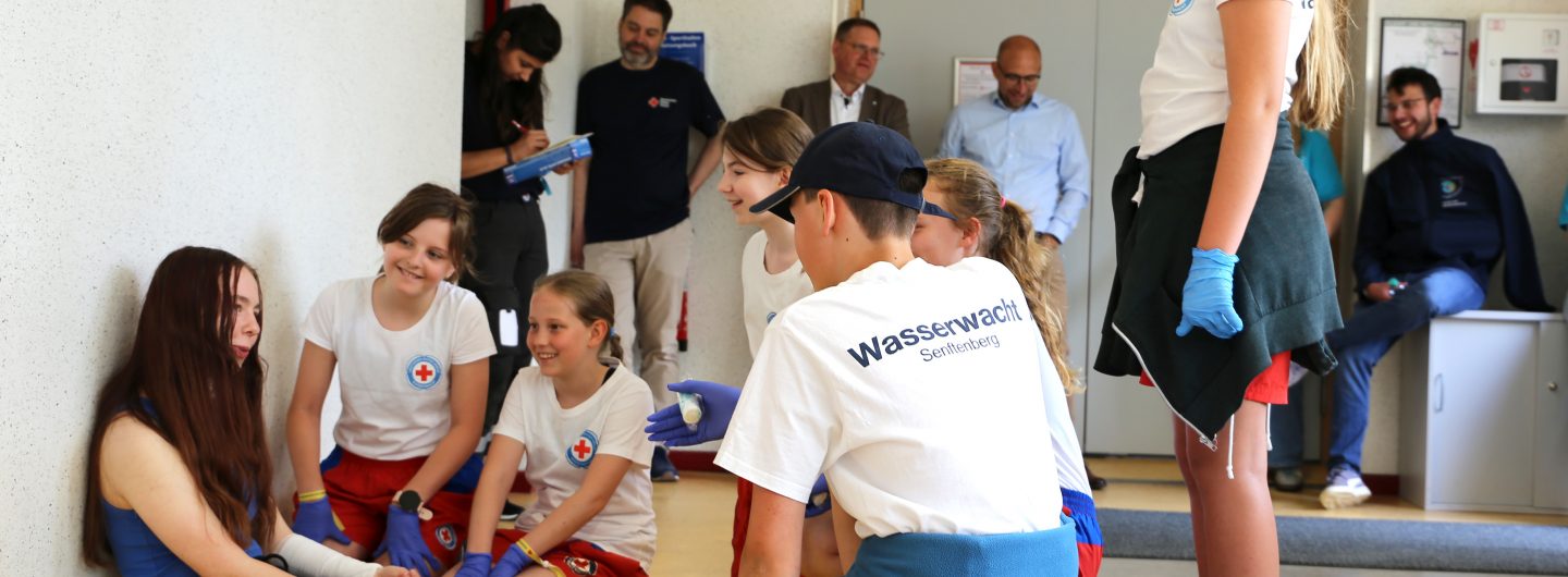 Fünf Kinder in T-Shirts der Wasserwacht des DRK kümmern sich in einer inszenierten Szene um eine Verletzte.