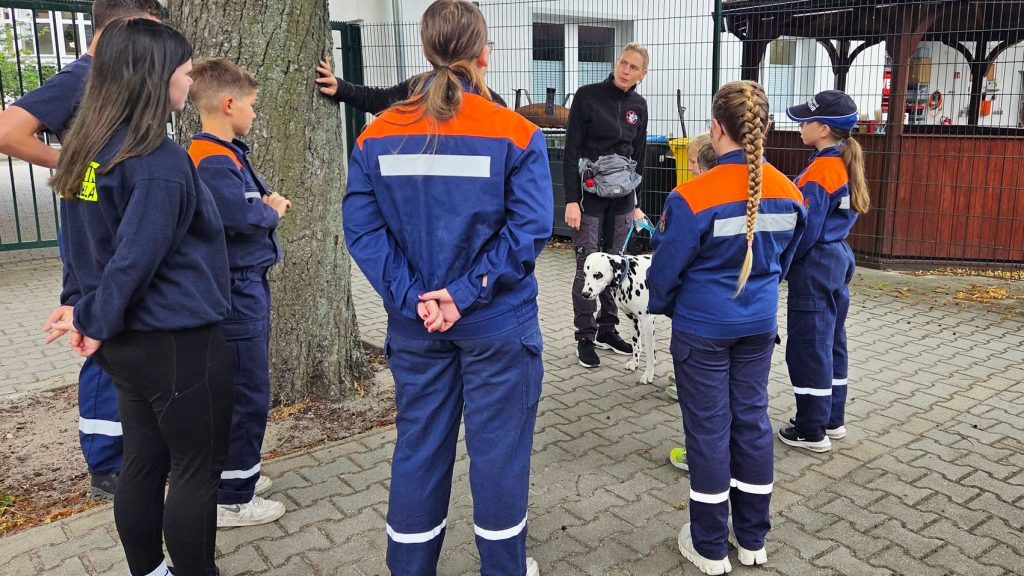 Kinder und Jugendliche in Feuerwehrausrüstung stehen im Halbkreis um eine erwachsene Person, die einen Dalmatiner an der Leine hält und offenbar etwas erklärt.
