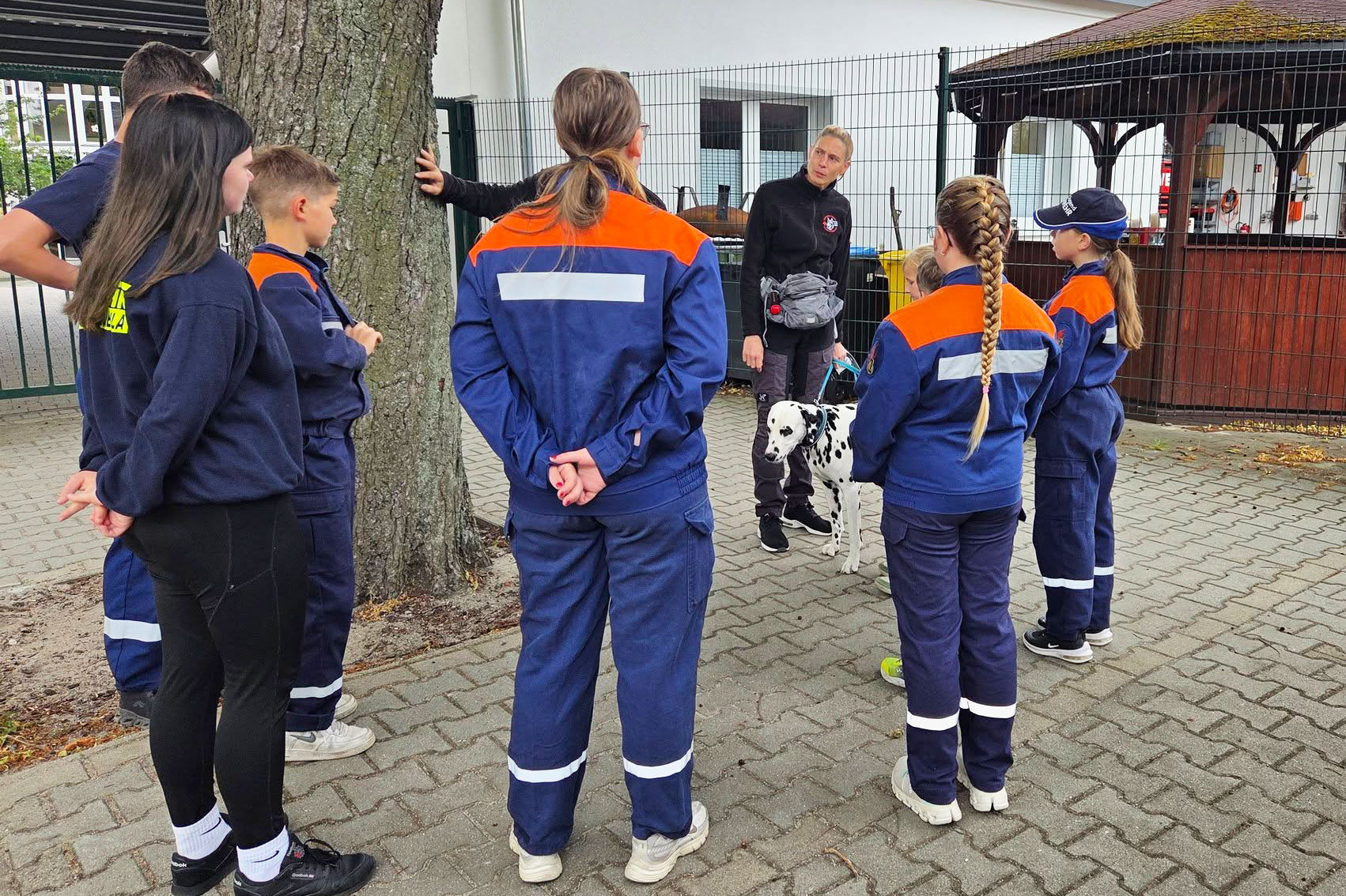 Kinder und Jugendliche in Feuerwehrausrüstung stehen im Halbkreis um eine erwachsene Person, die einen Dalmatiner an der Leine hält und offenbar etwas erklärt.