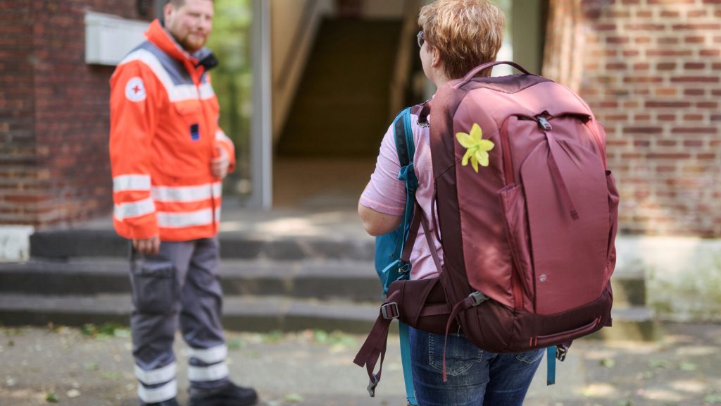 Eine Frau kommt mit zwei Rucksäcken bepackt in eine Notunterkunft und wird von einem Mann in Einsatzkleidung des DRK begrüßt.