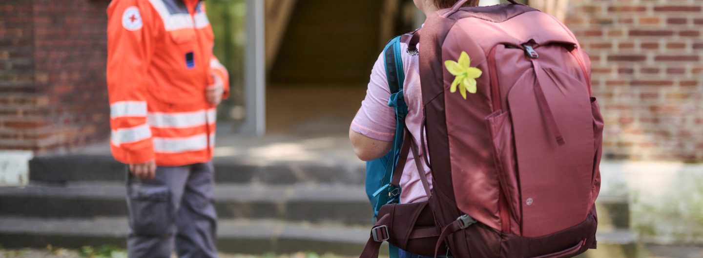 Eine Frau kommt mit zwei Rucksäcken bepackt in eine Notunterkunft und wird von einem Mann in Einsatzkleidung des DRK begrüßt.
