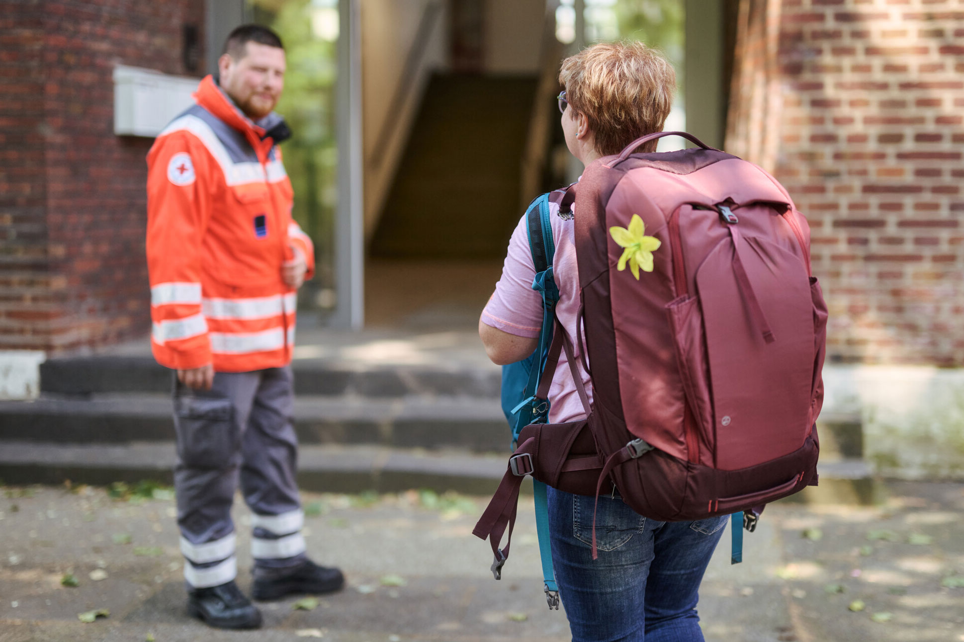 Eine Frau kommt mit zwei Rucksäcken bepackt in eine Notunterkunft und wird von einem Mann in Einsatzkleidung des DRK begrüßt.