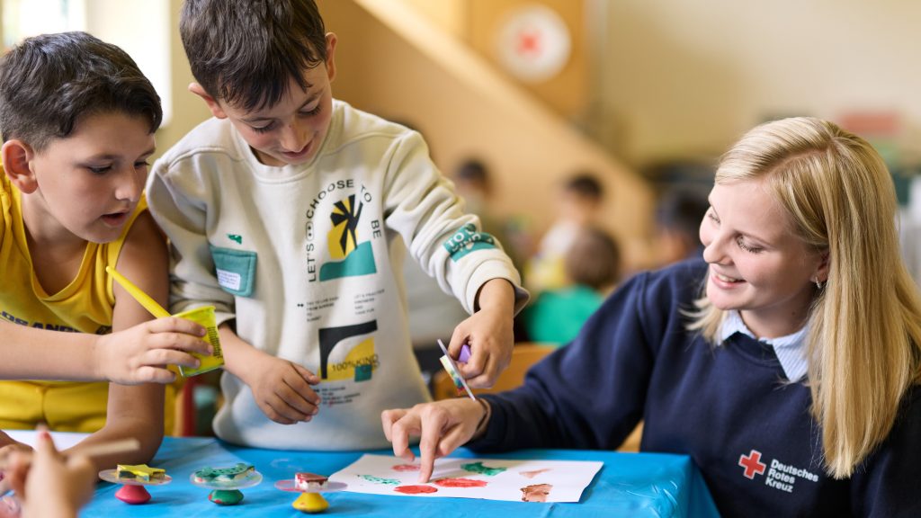 Eine junge Frau bastelt mit zwei Kindern an einem Tisch. Die Frau trägt einen Pulli mit einem Logo des Roten Kreuz