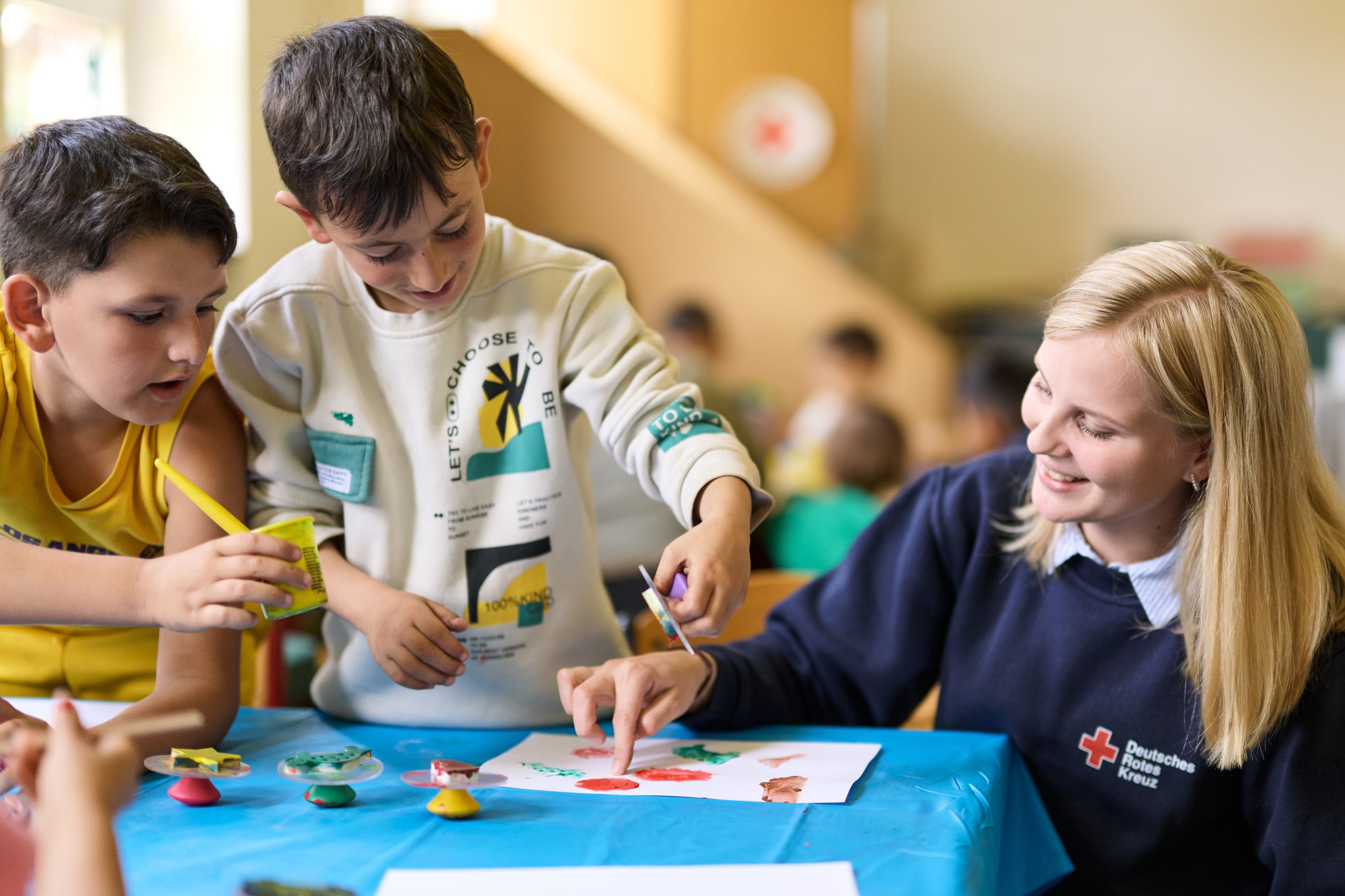 Eine junge Frau bastelt mit zwei Kindern an einem Tisch. Die Frau trägt einen Pulli mit einem Logo des Roten Kreuz
